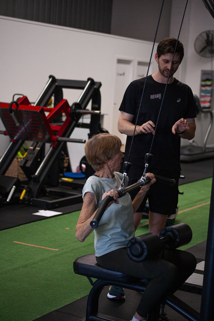 Person using a gym machine with another person assisting in a fitness setting