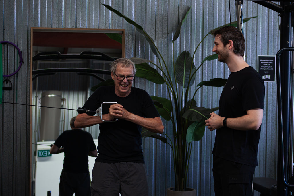 Two men in black shirts standing next to a large plant indoors. One physio and one client strength training.
